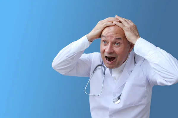 Доктор в белом халате с фонендоскопом на шее, крича от отчаяния, сжимая голову обеими руками на синем фоне — стоковое фото