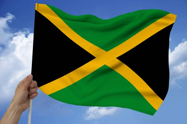 Mano masculina sostiene contra el fondo del cielo con nubes la bandera nacional de Jamaica en una textura lujosa de satén, seda con olas, primer plano, espacio de copia, concepto de viaje, economía, política — Foto de Stock