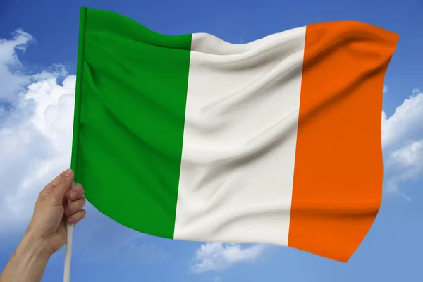 Χέρι κρατά στο φόντο του ουρανού με σύννεφα η έγχρωμη σημαία της Ιρλανδίας στην υφή του υφάσματος, μετάξι με κύματα, κοντινό — Φωτογραφία Αρχείου