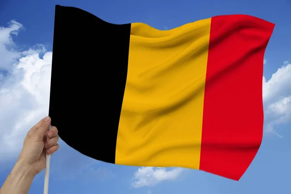 Hand holds against the background of the sky with clouds the colored flag of Belgium on the texture of the fabric, silk with waves, close-up — Stock Photo, Image