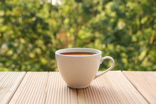 White cup with coffee on a light wooden table against a green summer garden, close-up, copy space — Stock Photo, Image