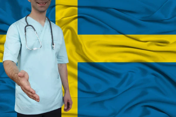 Male doctor in uniform professional clothes with a stethoscope welcomes patients with his hand against the background of the national flag, the concept of the country's health and medical insurance — Stock Photo, Image