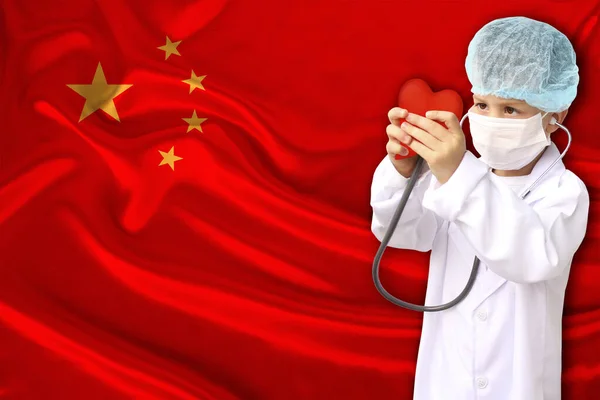 Фото дитини з стетоскопом у професійній формі з серцем у руках на задньому плані національного прапора Китаю, поняття педіатрії, хірургії, кардіології — стокове фото