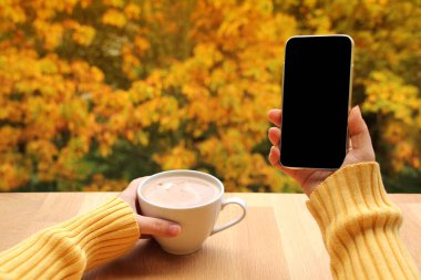 Bir elinde süt veya kapuçino olan bir fincan kakao ve Gargen 'de boş ekran cep telefonu olan akıllı telefon, sonbahar havası konsepti, yakın plan, uzay kopyası.