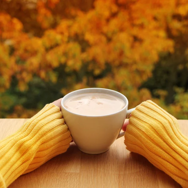 cup of cocoa with milk or cappuccino in the hands of a girl in a yellow sweater, outdoors with a view of yellow and orange trees, the concept of autumn mood, close-up