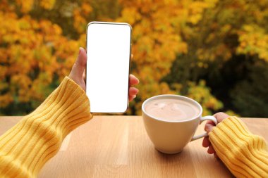 Bir elinde süt ya da kapuçino olan bir fincan kakao ve diğer elinde boş ekran olan bir akıllı telefon. Arka planda sarı ve turuncu ağaç yaprakları, sonbahar havası kavramı, yakın plan, boşluğu kopyalamak.