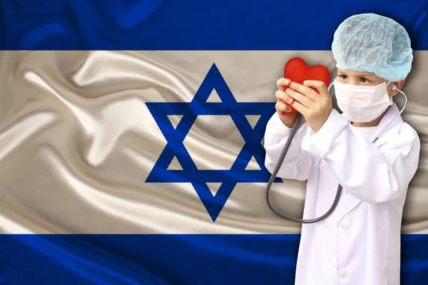 Child, boy, in a white doctor’s coat, hat and mask attached a stethoscope to a red heart model, Israel flag background, close-up, focus on the face, medical concept, cardiology, copy space — Φωτογραφία Αρχείου