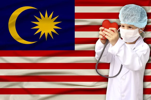 Child, boy, in a white doctor’s coat, hat and mask attached a stethoscope to a red heart model, Malaysia flag background, close-up, face focus, medical, cardiology concept, copy space — Stock Photo, Image