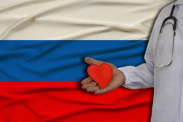 Foto de un médico con un estetoscopio con un corazón en la mano en el fondo de la bandera nacional del estado de la Federación de Rusia, el concepto de atención médica, tratamiento cardiológico — Foto de Stock