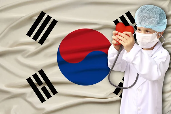 Child, boy, in a white doctor’s coat, hat and mask attached a stethoscope to a red heart model, South Korea flag background, close-up, face focus, medical, cardiology concept, copy space — Stock Photo, Image