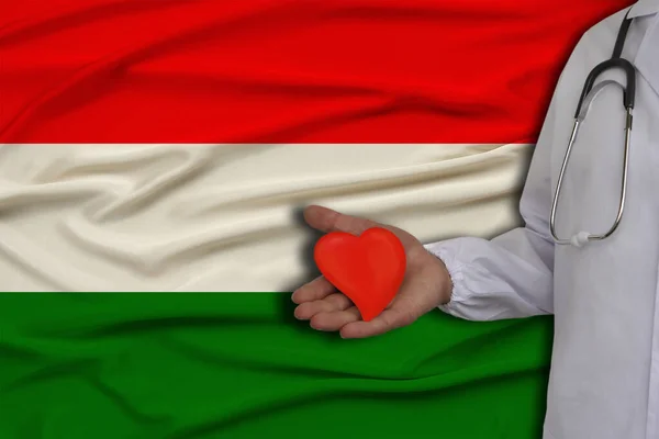 photo of a doctor with a stethoscope with a heart in his hand against the background of the national flag of the state of Hungary, the concept of health care, cardiological treatment, medical insuranc