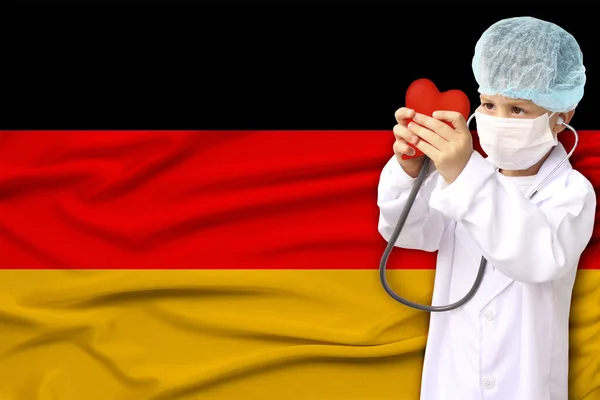 Дитина, хлопчик, в білому лікарському пальто, капелюх і маска прикріпили стетоскоп до червоної моделі серця, фон прапора Німеччини, зблизька, фокус на обличчі, медична концепція, кардіологія, копіювальний простір — стокове фото