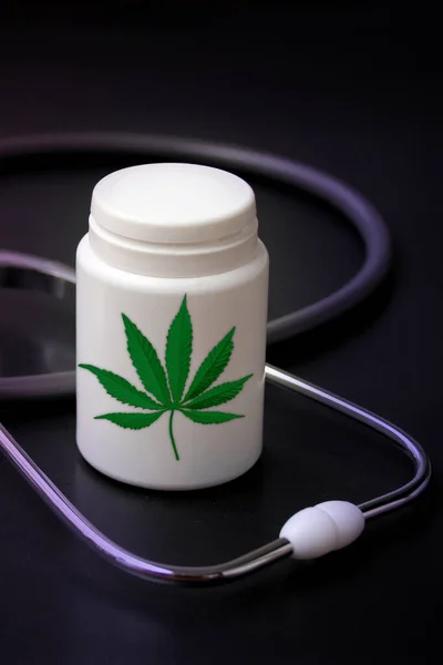 Estetoscopio médico se encuentra en un fondo negro, un frasco blanco de medicina con un icono de la hoja de marihuana, concepto de cannabis medicinal, sustancias narcóticas, primer plano, espacio de copia —  Fotos de Stock