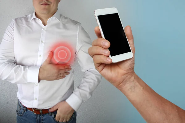 Мужчина в белой рубашке держится за грудь в области сердца, рядом с его рукой держит телефон с черным дисплеем, концепция проблем со здоровьем, вызова врача, скорой помощи, копирования пространства — стоковое фото