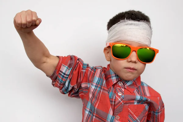 Ragazzo in occhiali da sole scuri, in camicia a quadri, la testa fasciata, controlla i muscoli, il concetto di pazienza dei bambini, primo piano — Foto Stock