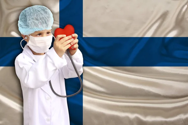 Barn, pojke, i en vit läkare päls, hatt och mask fäst ett stetoskop till en röd hjärtmodell, Finland flagga bakgrund, närbild, fokus på ansiktet, medicinska begrepp, kardiologi, kopia utrymme — Stockfoto