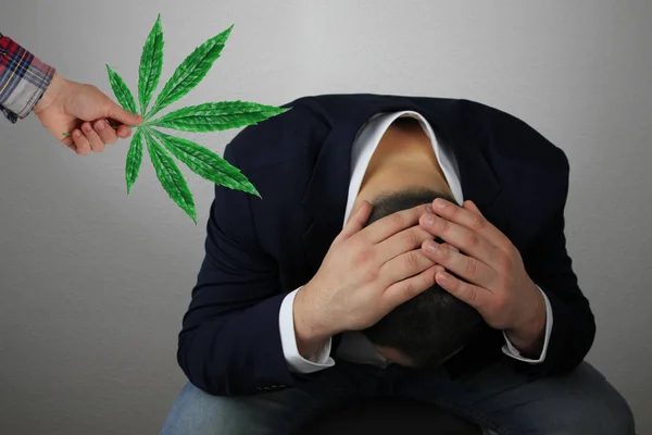 Mano sostiene hoja verde de marihuana al hombre de negocios sentado apretando la cabeza en las manos, símbolo de problemas, dolor, concepto de cannabis medicinal, drogas narcóticas, primer plano —  Fotos de Stock