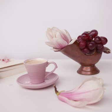 Beyaz bir masada bir fincan kapuçino, çiçek açan manolya tomurcukları, pembe arka planda beyaz bir kapakta bir kitap, tasarımcı için bir boşluk, bir davetiye formu, bahar havası konsepti.