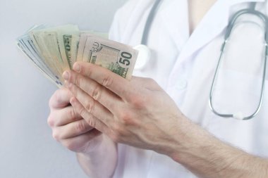 Koruyucu maskeli bir erkek doktor, elinde steteskopla elinde dolar banknotları, sağlık sigortası kavramı, bütçe, tedavi masrafları, organ bağışı hayranı.