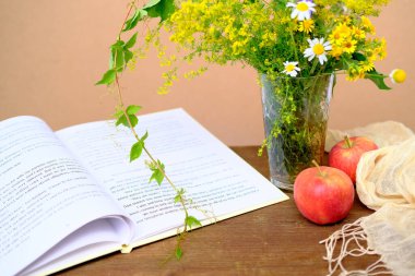 Tahta bir masanın üzerinde açık bir kitap, kırmızı elmalar, bir buket kır çiçeği, yaz havası, hobiler, doğal yiyecekler.