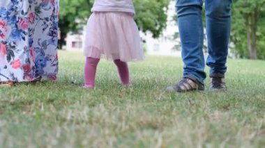 Uzun elbiseli bir kadın, anne, sana küçük bir çocuğu gezdirmeyi öğretiyor, yazın çimenlerin üzerinde sarı saçlı bir kız, aile yaz tatili konsepti, açık hava oyunları, ilk adımlar, çocuk gelişimi.