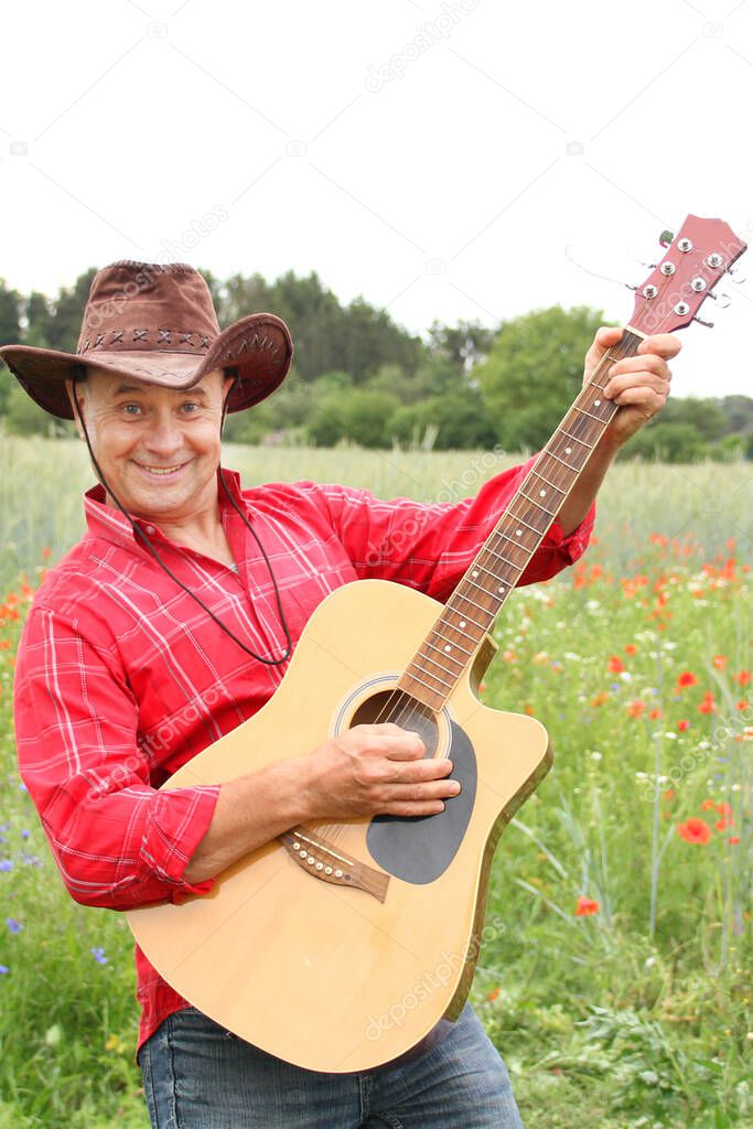 cheerful adult man in a red shirt and a coboy hat on a field of green poppies plays guitar, posing and having fun, concept musical, inspiration by nature, beauty of the native land