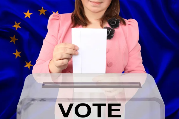 woman voter in smart pink jacket lowers the ballot in a transparent ballot box against the background of the European Union national flag, concept of state elections, referendum