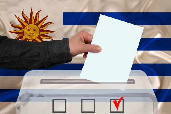 男性の有権者は ウルグアイの国旗 州の選挙の概念 国民投票の背景にある透明な投票箱に投票を落とします — ストック写真