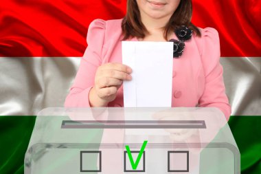 Kadın seçmen, Macaristan bayrağı, devlet seçimleri ve referandumun arka planına karşı şeffaf bir oy sandığını indiriyor