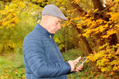 Gri şapkalı, mavi ceketli, sonbahar parkında sarı ve turuncu ağaçların arasında dikilen yaşlı bir adamın renkli fotoğrafı telefon ediyor.