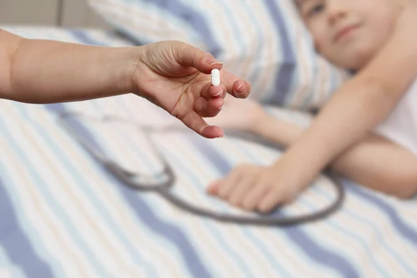 female hand holds out, offering, white elongated pill, close-up, blurred image of a lying child in the background, copy space, concept of medical care, treatment