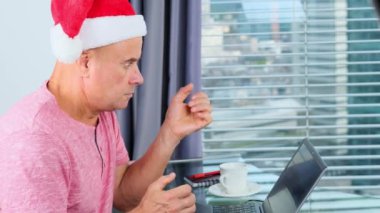 Noel Baba şapkalı yetişkin bir adam dizüstü bilgisayarın önünde oturur, bilgi alır ve duygusal tepki verir, Noel konsepti, yeni yıl kutlaması, müdire, Noel 'i bekler, Noel' i bekler,