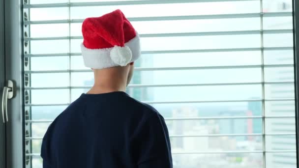 头戴圣诞礼帽的年轻人站在窗前 在窗后看得见欧洲的大城市 他们的旅游观念 新年庆祝活动 西尔维斯节 等待节日 圣诞节销售 — 图库视频影像