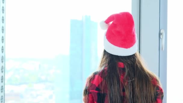 サンタ クラスの帽子の若い女性が窓の前に立っています その後ろにはヨーロッパの大都市 新年のお祝い シルベスター 休日を待っています クリスマスの販売 — ストック動画