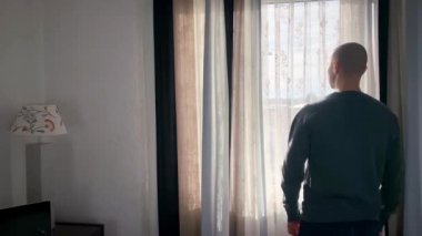 Genç adam pencerenin önünde duruyor, perdeleri açıyor, sokağa bakıyor, iş seyahati konsepti, turizm, yalnızlık, evde kalmak, karantina