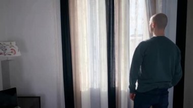 Genç adam pencerenin önünde duruyor, perdeleri açıyor, sokağa bakıyor, iş seyahati konsepti, turizm, yalnızlık, evde kalmak, karantina