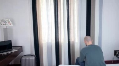 Genç adam pencerenin önünde duruyor, perdeleri açıyor, sokağa bakıyor, iş seyahati konsepti, turizm, yalnızlık, evde kalmak, karantina