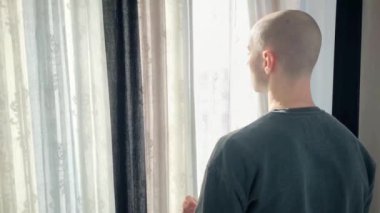 Genç kel adam pencerenin önünde duruyor, perdeleri açıyor, sokağa bakıyor, iş seyahati konsepti, turizm, yalnızlık, evde kalmak, karantina