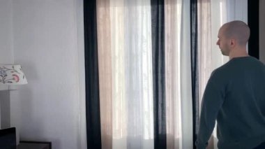 Genç kel adam pencerenin önünde duruyor, perdeleri açıyor, sokağa bakıyor, iş seyahati konsepti, turizm, yalnızlık, evde kalmak, karantina