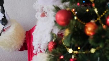 Kırmızı takım elbiseli yetişkin Noel Baba masaya oturur, Noel ağacı toplar, çelenkler, Noel konsepti, yeni yıl kutlaması, tatil satışları ve indirimlerle süslenir.