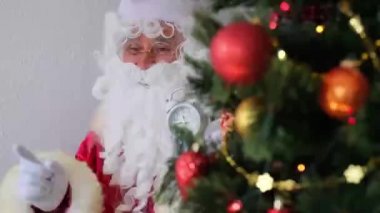Kırmızı takım elbiseli Noel Baba masaya oturur, çalar saati gösterir, Noel ağacı toplarla süslenir, Noel konsepti, yeni yıl kutlaması, tatil satışları... 