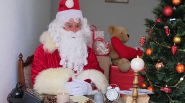 Kırmızı takım elbiseli yetişkin Noel Baba masaya oturur, Noel ağacı toplar, çelenkler, Noel konsepti, yeni yıl kutlaması, tatil satışları ve indirimlerle süslenir.