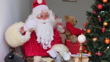 Kırmızı takım elbiseli yetişkin Noel Baba masaya oturur, Noel ağacı toplar, çelenkler, noel konsepti, yeni yıl kutlaması, tatil satışları ve indirimler ile güzelce süslenir.