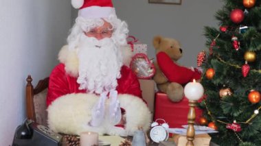 Kırmızı takım elbiseli yetişkin Noel Baba masaya oturur, Noel ağacı toplar, çelenkler, Noel konsepti, yeni yıl kutlaması, tatil satışları ve indirimlerle süslenir.