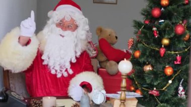 Kırmızı takım elbiseli yetişkin Noel Baba masaya oturur, Noel ağacı toplar, çelenkler, Noel konsepti, yeni yıl kutlaması, tatil satışları ve indirimlerle süslenir.