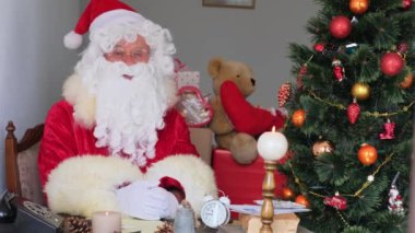 Kırmızı takım elbiseli yetişkin Noel Baba masaya oturur, Noel ağacı toplar, çelenkler, Noel konsepti, yeni yıl kutlaması, tatil satışları ve indirimlerle süslenir.
