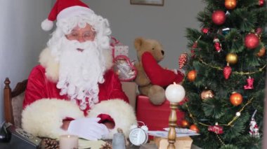Kırmızı takım elbiseli yetişkin Noel Baba masaya oturur, Noel ağacı toplar, çelenkler, Noel konsepti, yeni yıl kutlaması, tatil satışları ve indirimlerle süslenir.