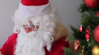 Kırmızı takım elbiseli beyaz sakallı Noel Baba toplarla süslenmiş bir Noel ağacının yanında oturuyor, kalemle bir mektup yazıyor, Noel konsepti, hediyeleri bekliyor, kutluyor.
