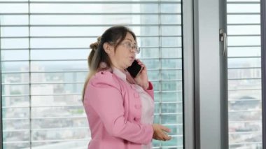 Pembe elbiseli yetişkin bir kadın modern bir Avrupa şehrinin ofisinde büyük bir pencerenin önünde duruyor, cep telefonuyla konuşuyor, ilişki kavramı, iletişim