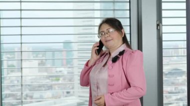 Pembe elbiseli yetişkin bir kadın modern bir Avrupa şehrinin ofisinde büyük bir pencerenin önünde duruyor, cep telefonuyla konuşuyor, ilişki kavramı, iletişim